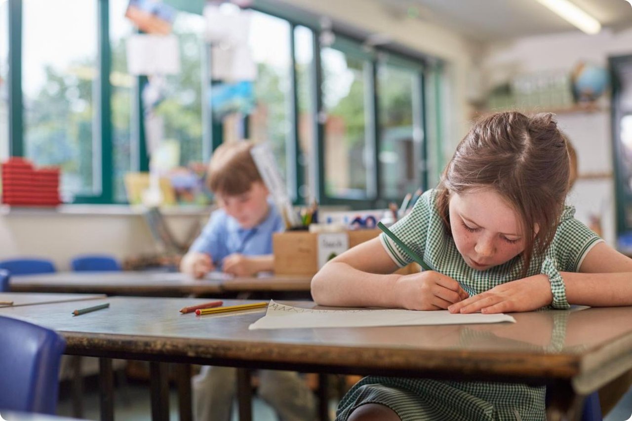 Primary school girl in a green checked school uniform summer dress writing attentively on a piece of paper on the desk in front of her.