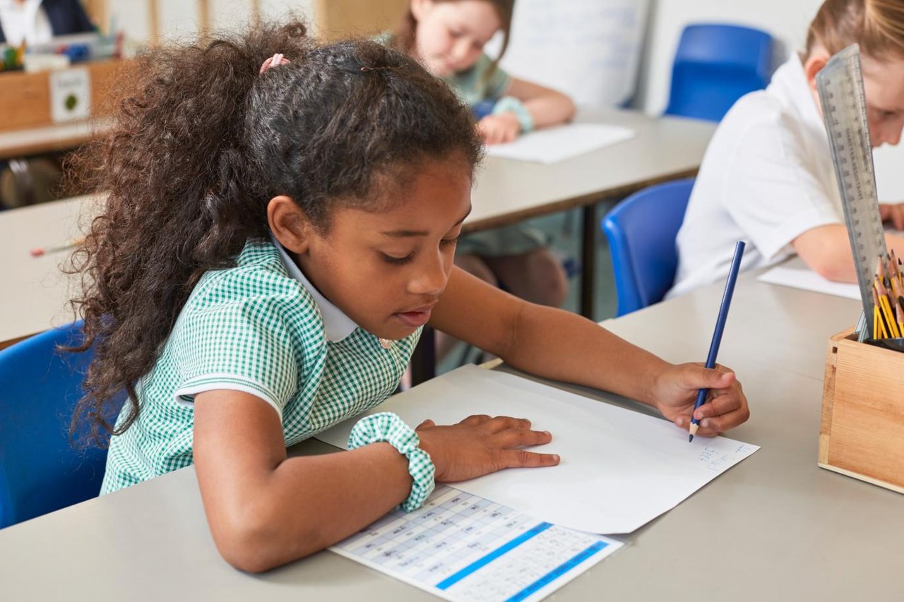 Primary school girl wearing green and white checked summer dress, working studiously at a desk with papers in front of her