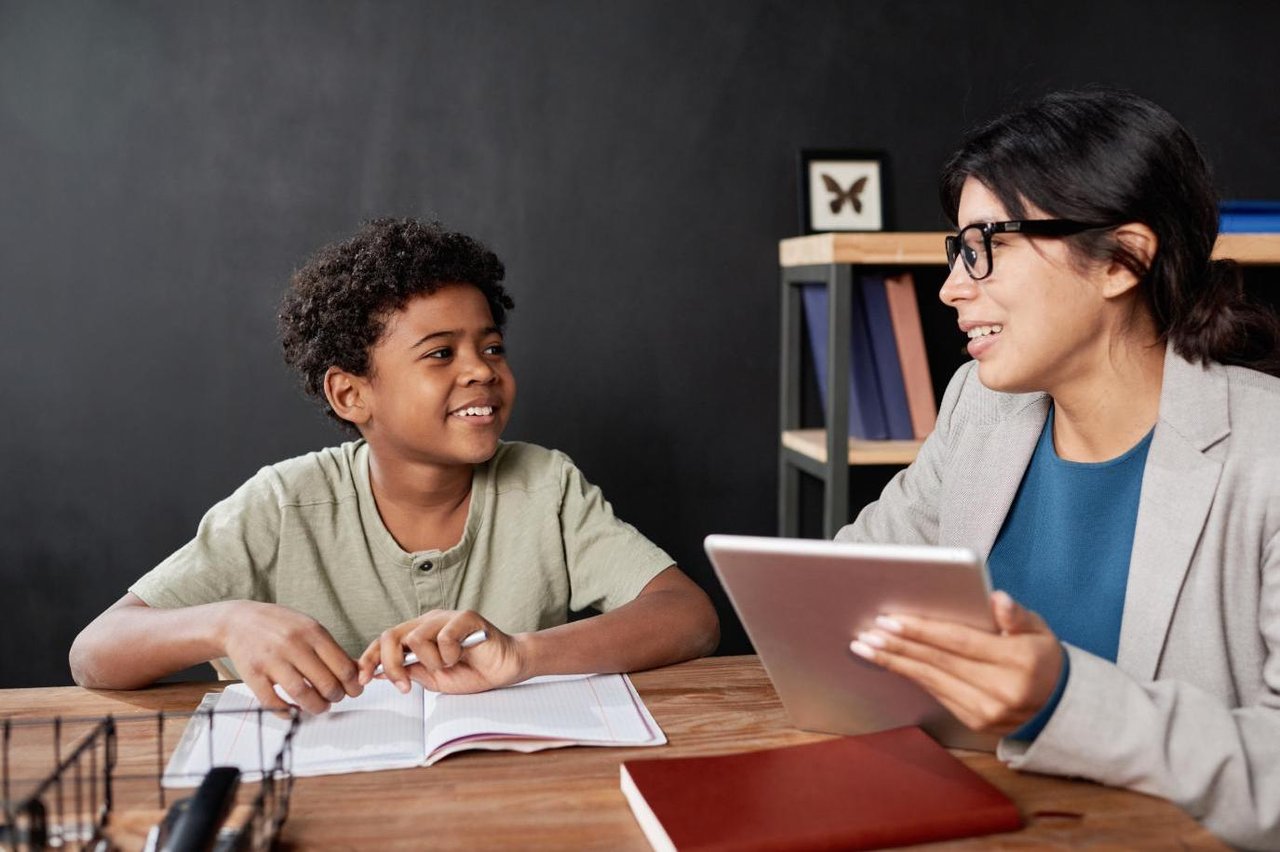 Young boy and adult female teacher sitting at a table and smiling at each other. The boy has an open notebook in front of him and the teacher is holding a digital tablet.