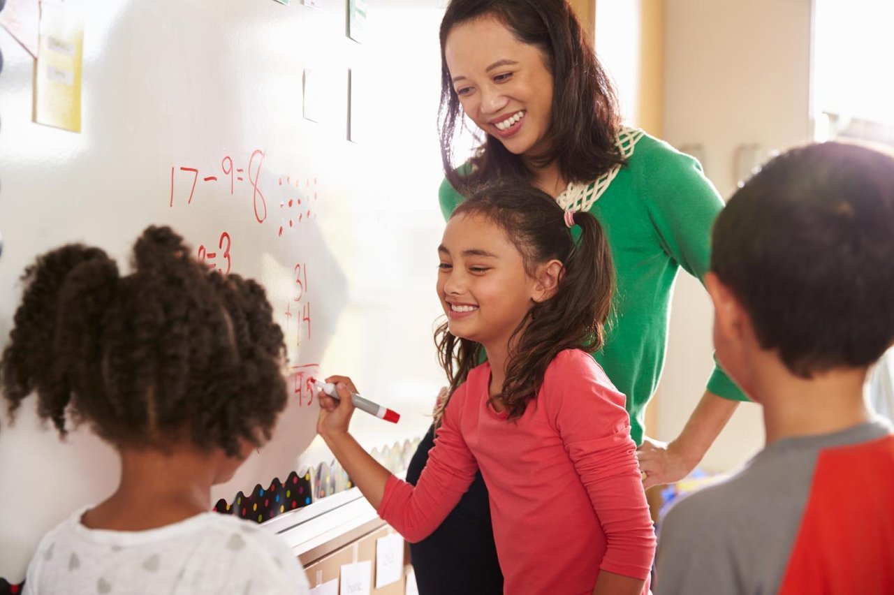 Young schoolgirl and teacher smiling while writing mathematical sums on the classroom whiteboard