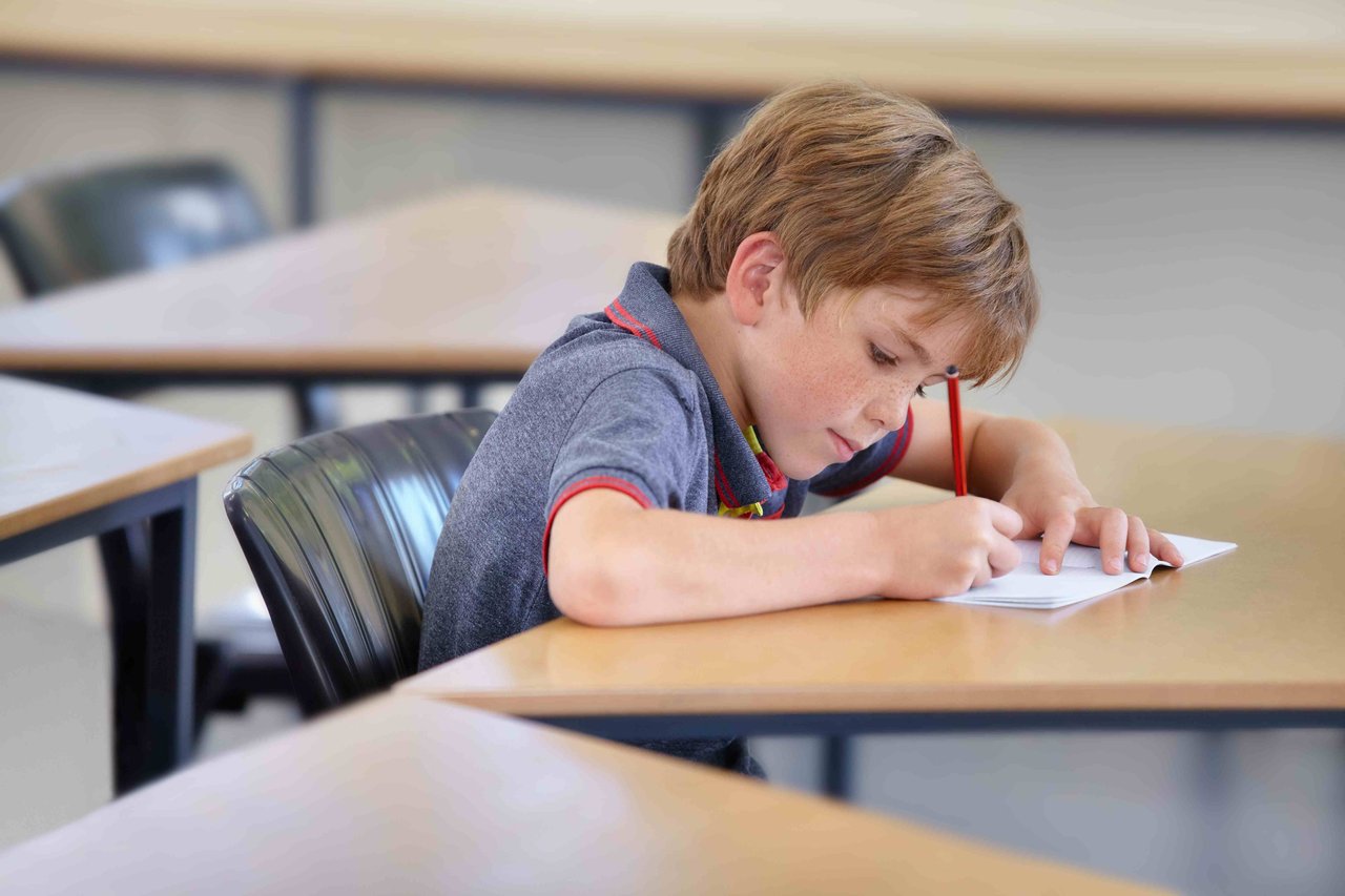 Young schoolboy in grey school shirt with red hems, sitting at a school desk in an empty classroom writing in an open workbook.