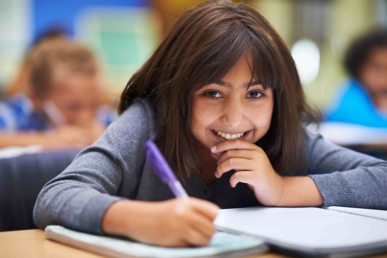 Young girl in a long-sleeved grey top writing in a notebook with a purple pen, smiling at the camera. There are other children in the background behind her.