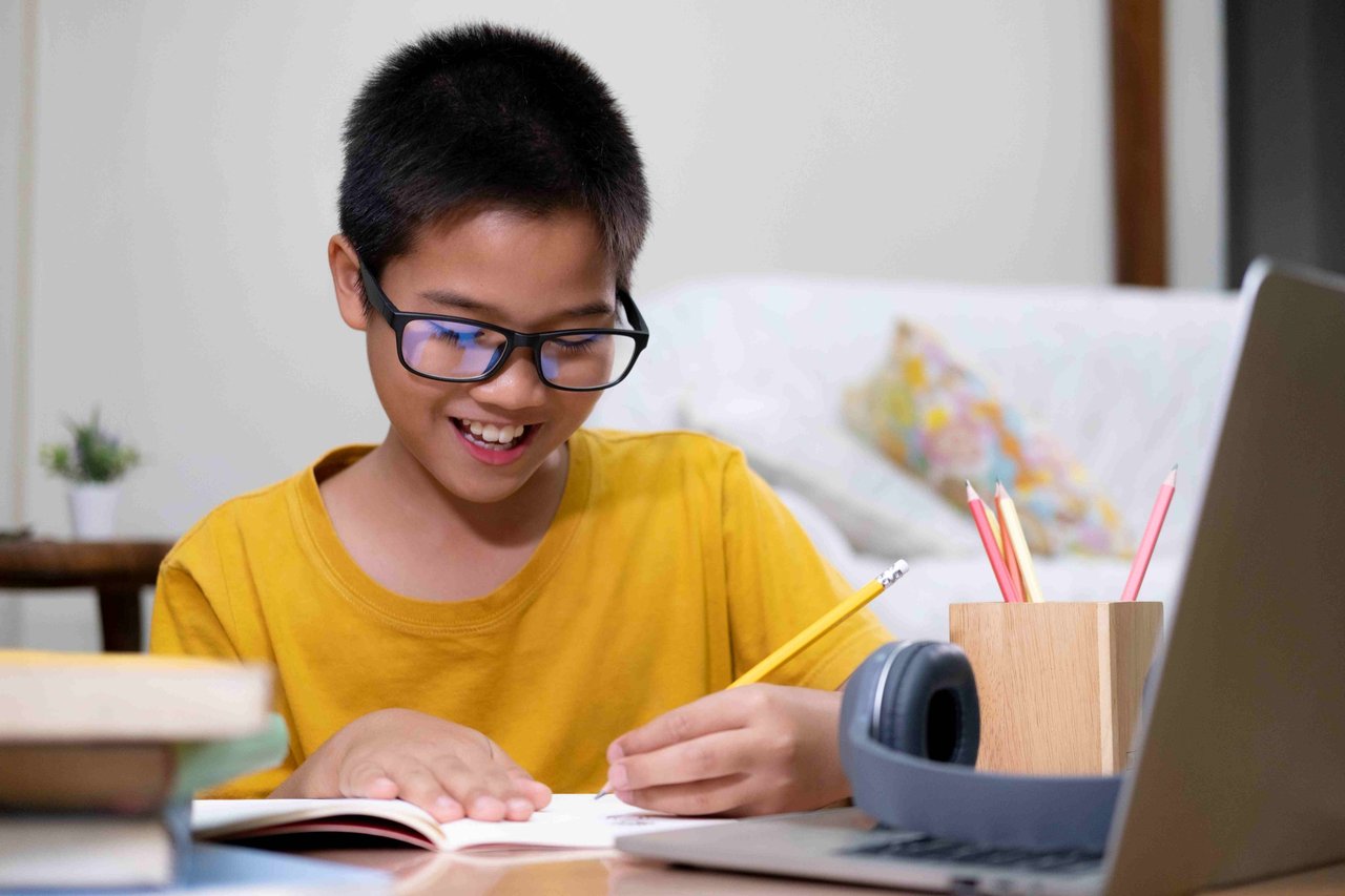 Young boy wearing glasses and a yellow t-shirt smiling while writing with a pencil in a notebook