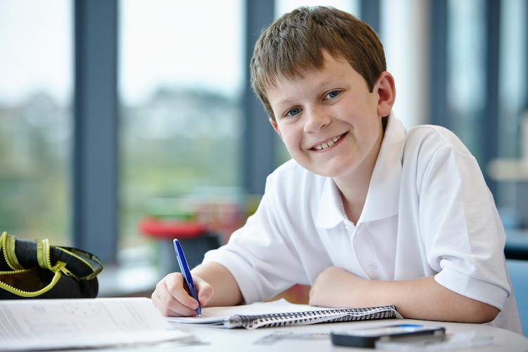 A smiling boy with brown hair, writing on a notepad in a school classroom