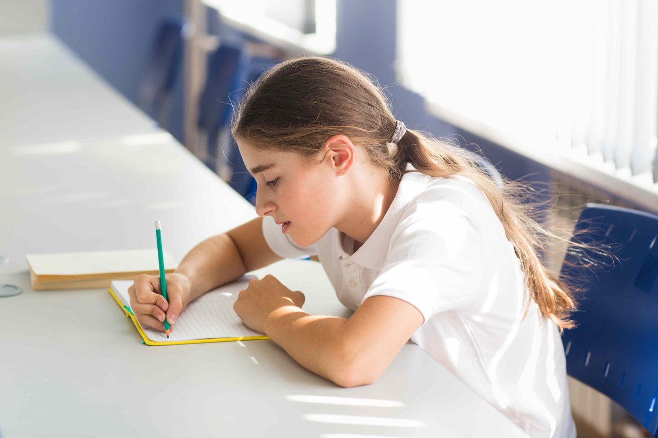 Schoolgirl with a long ponytail in a white polo top sitting at a desk in an empty classroom, studiously writing with a pencil in a notebook