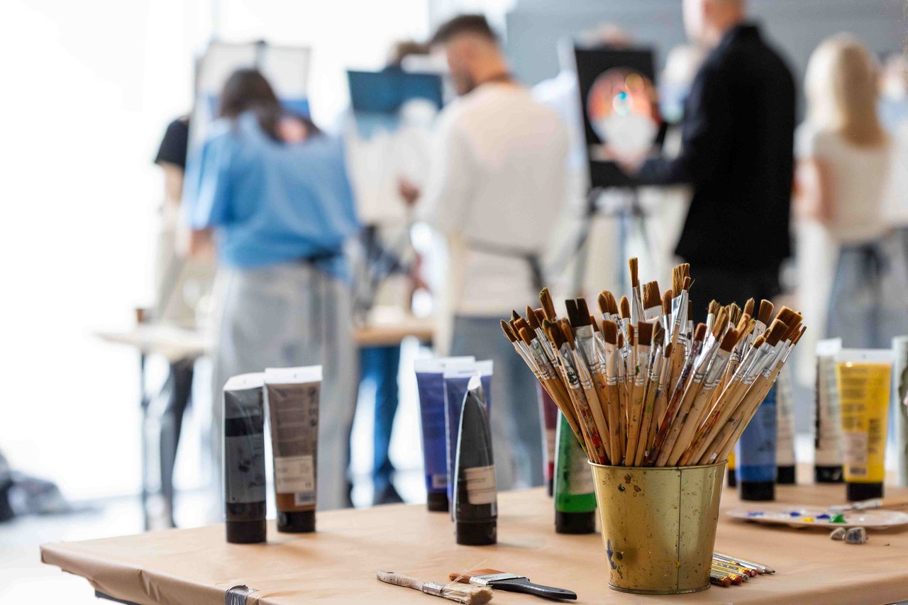 A photograph of some paint brushes and paint with a group of students painting in the backgroun