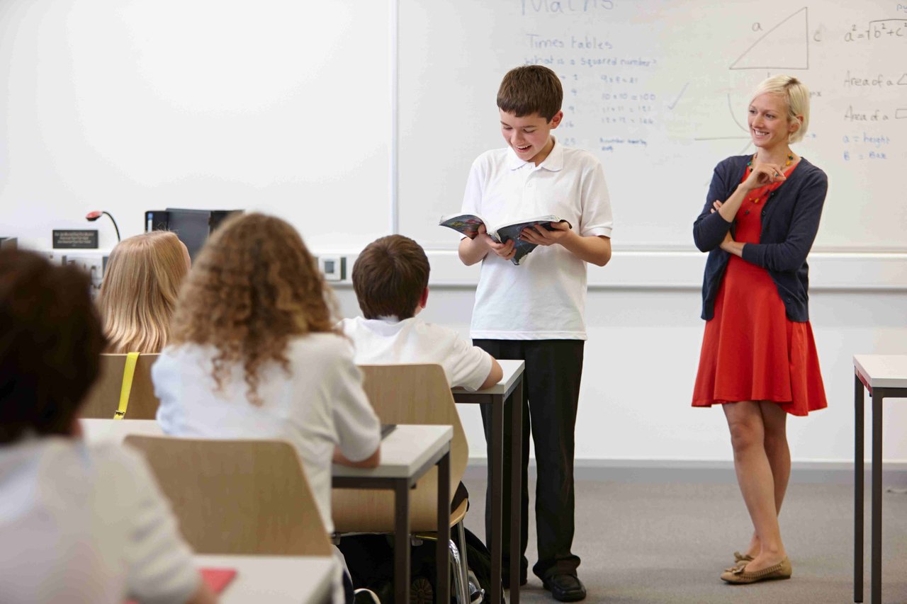 A school boy stood in front a classroom full of students reading from a book, with a female teacher stood by his side