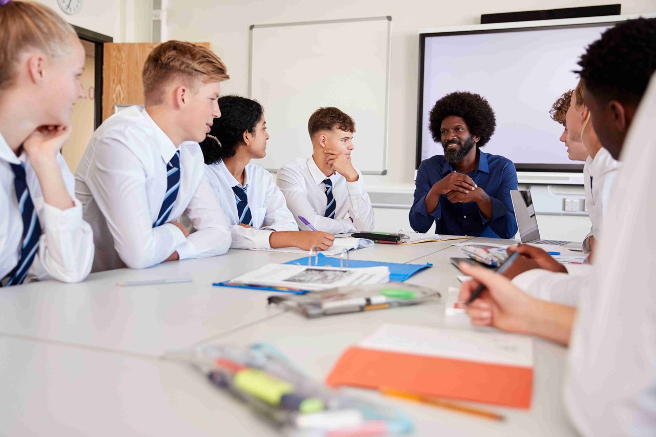 A group of students in uniform facing towards a teacher, all sat around a classroom table