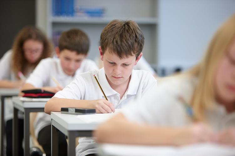 A young boy with brown hair wearing a white polo top, writing an exam with a pencil