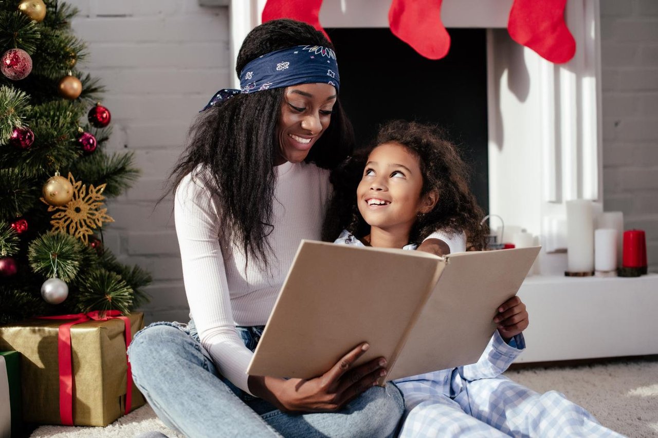 Mother and child sitting on the floor reading a book together. A Christmas tree is pictured in the corner.