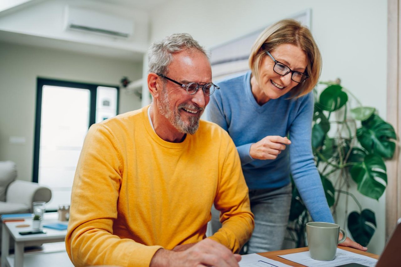 Middle-aged man in glasses and a yellow jumper, and a middle-aged woman in glasses and a blue jumper, smiling while looking at a laptop screen