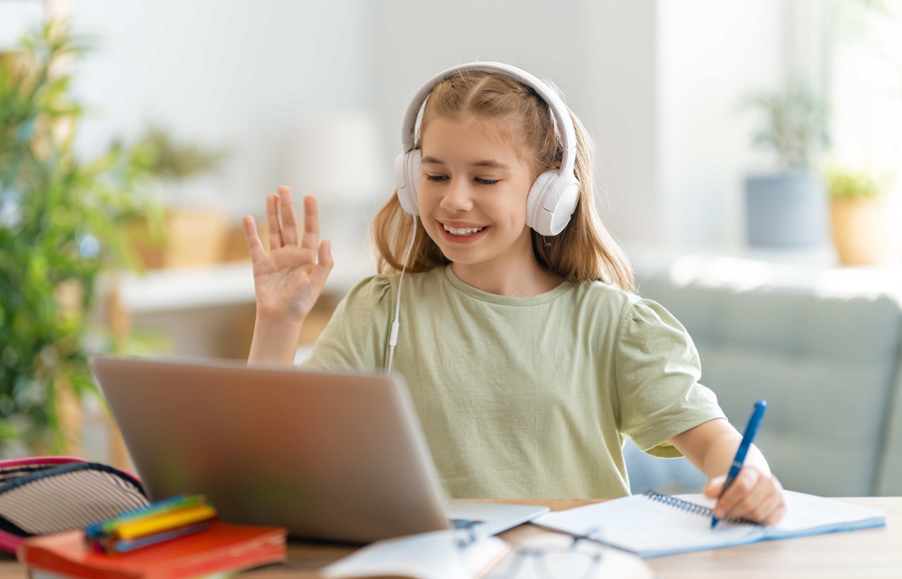  Blonde girl wearing a light green t-shirt and white headphones, sitting in a bright and modern room and waving at someone on a laptop
