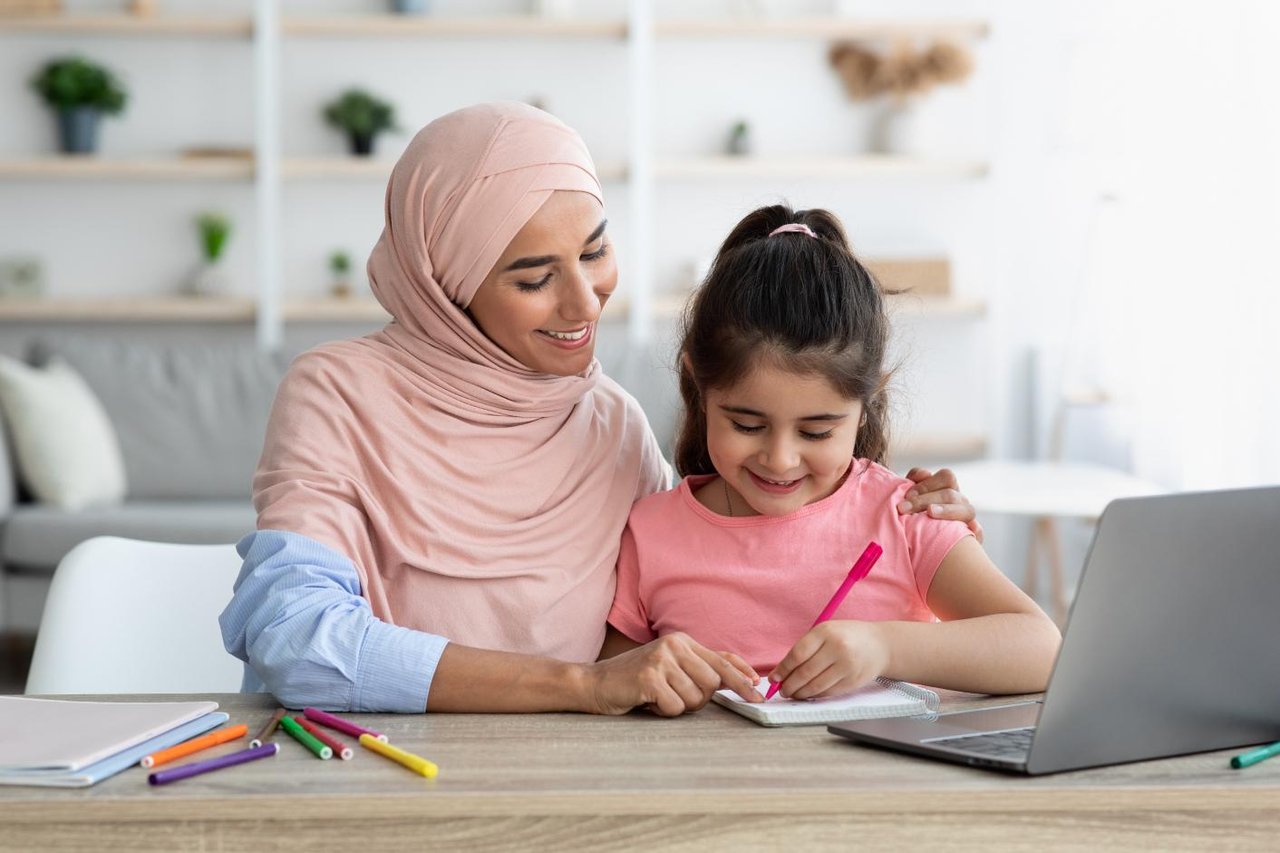 Mother sitting at a table with her arm around her daughter, who is writing in a notebook. Both mother and daughter are smiling. 