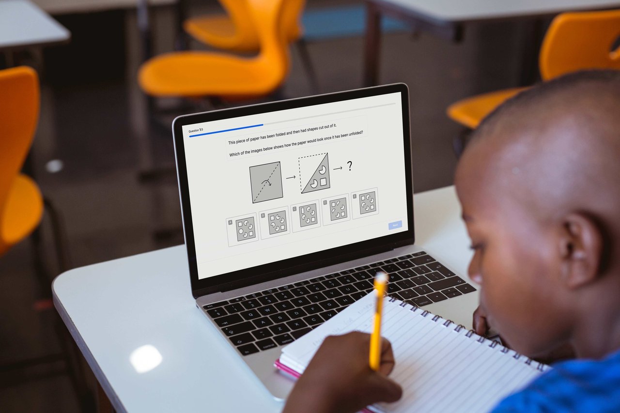 Boy sitting at a school desk in a classroom in front of a laptop with a Quest non-verbal reasoning question on the screen. He is holding a pencil and writing in a lined notebook.