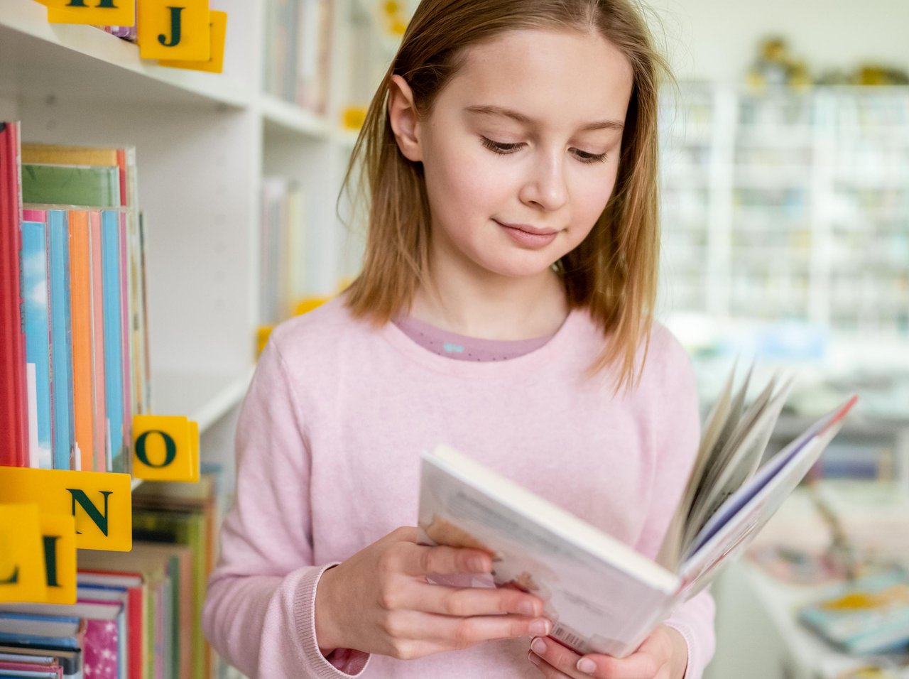 Young girl in a pale pink jumper standing next to a library bookshelf, looking at an open book in her hands