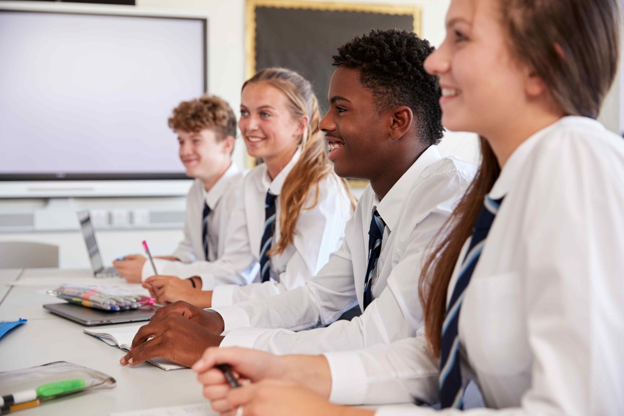 Four teenagers (two boys and two girls) wearing white school shirts and blue striped school ties, sitting next to each other at a table in a school classroom and smiling at something off-camera.