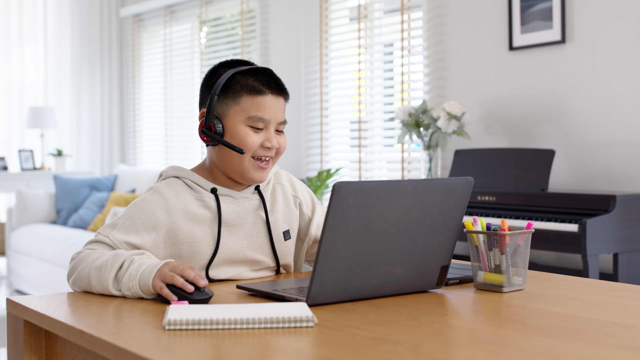 Young Asian boy wearing a headset smiling at someone not shown on a laptop, while sitting at a wooden table in a bright and modern family living room.