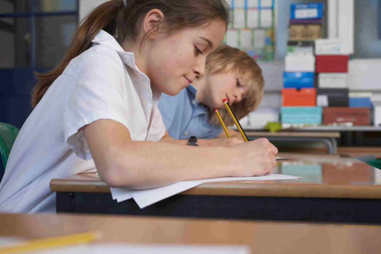 Girl in a white school shirt writing with a pencil on paper at a school desk. A boy in a school shirt is also writing on a piece of paper in the background.