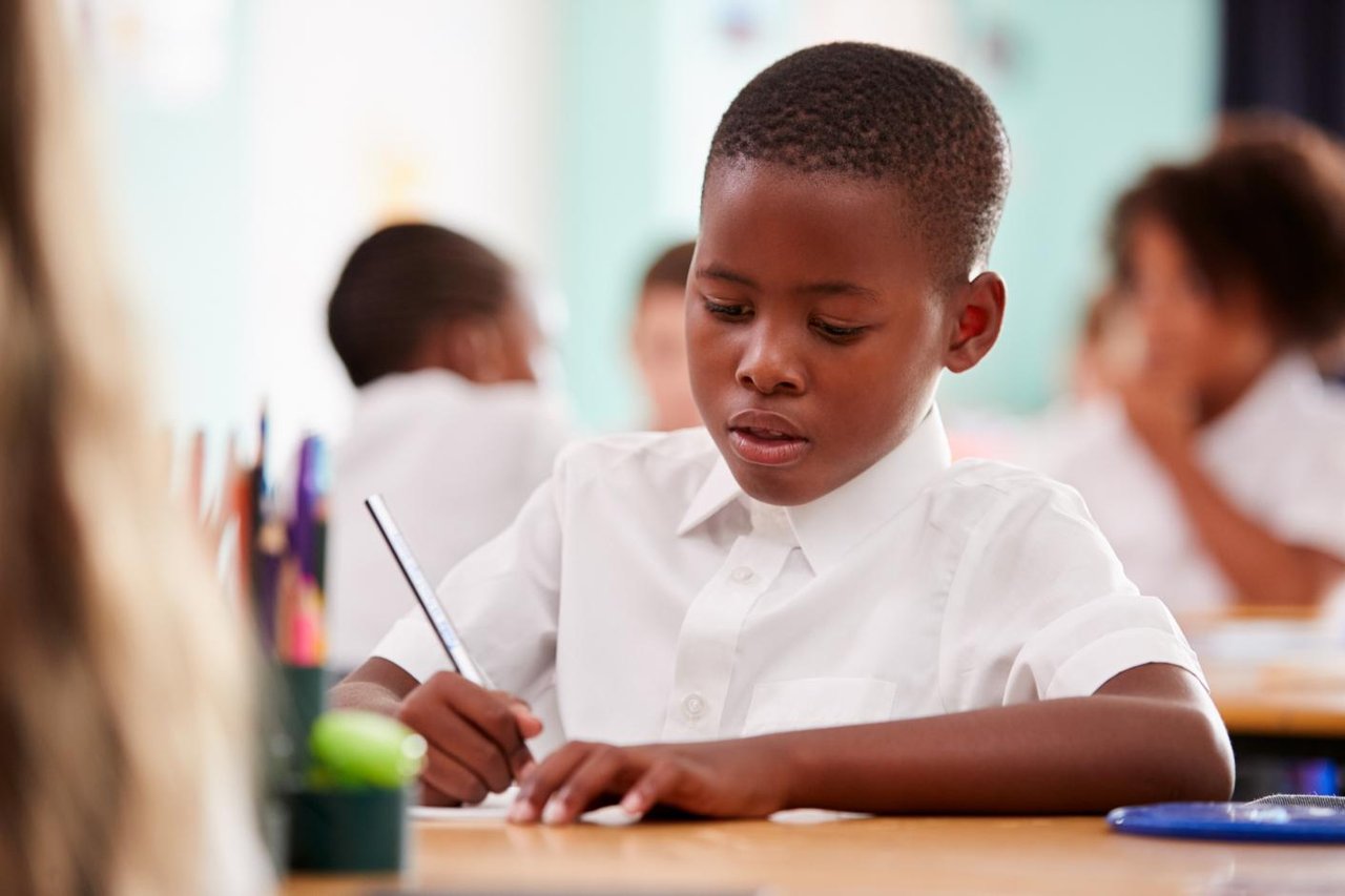 Young schoolboy in buttoned up white short-sleeve shirt writing at a desk in a classroom with other students in the background