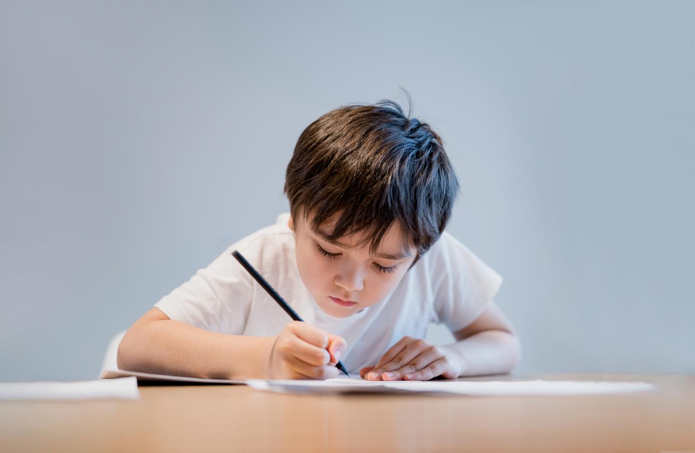 Young boy in a white t-shirt concentrating while writing on a piece of paper at a table