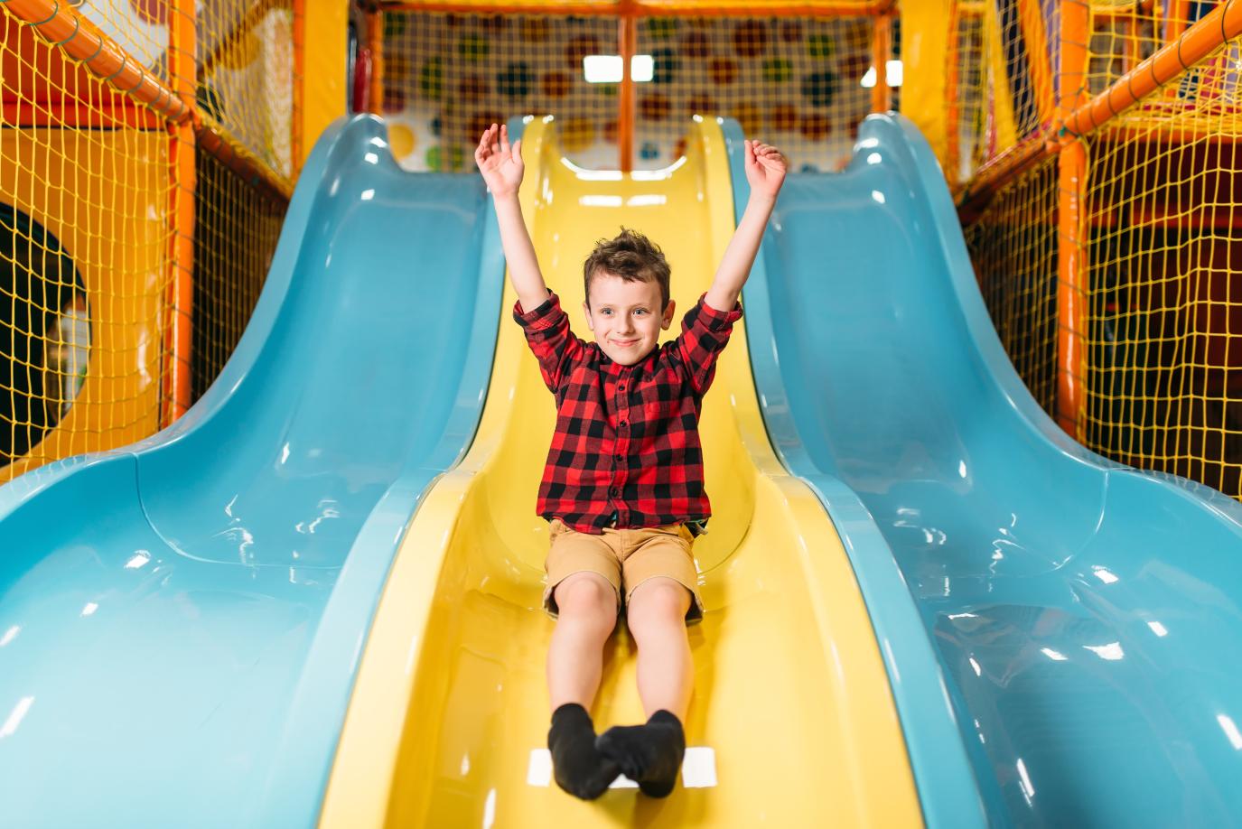 Young smiling boy sliding down an indoor slide with his arms in the air