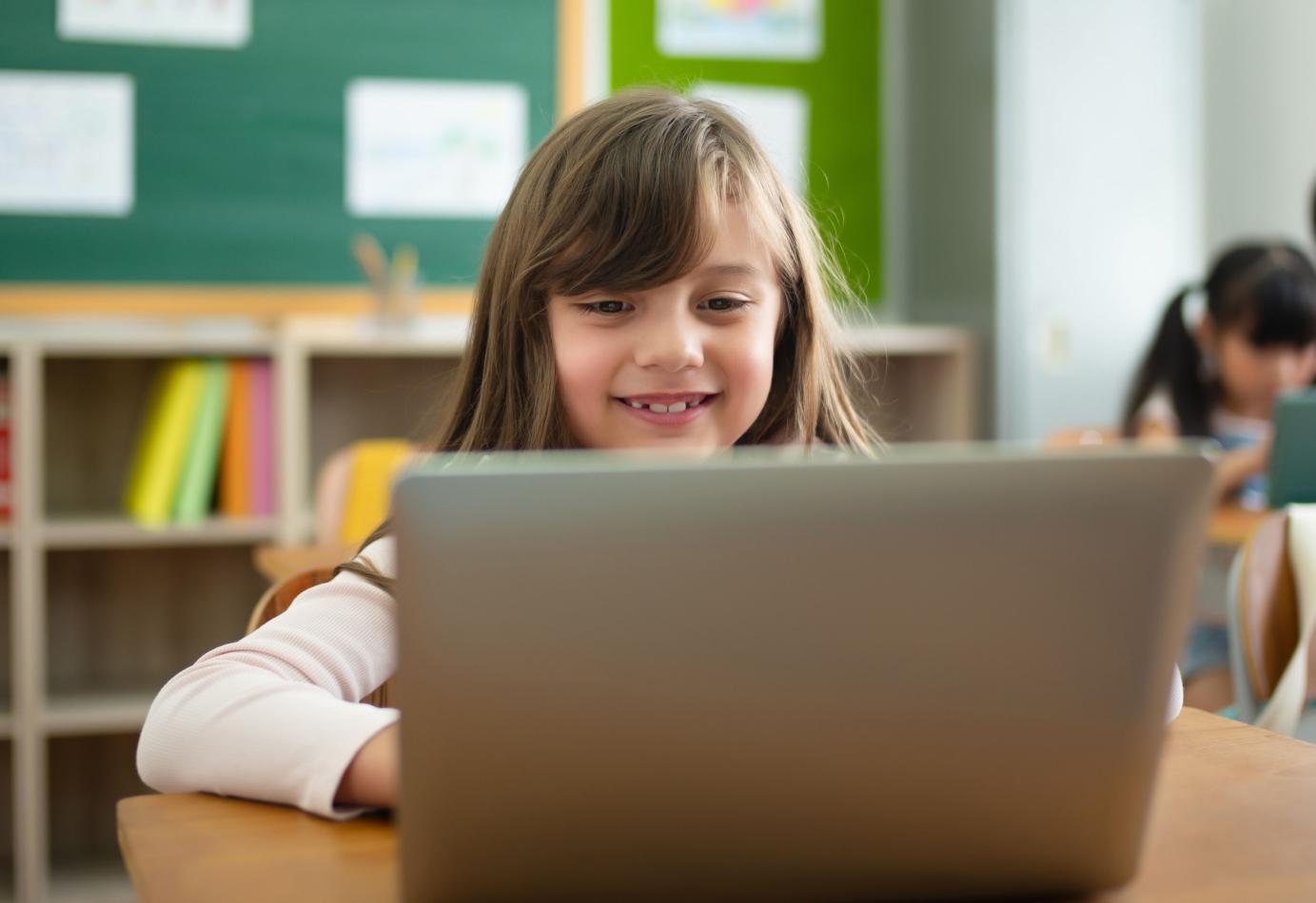 Young girl in a school classroom, smiling while looking at something on a laptop screen