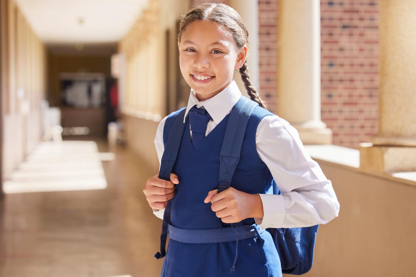 Girl with plaited hair wearing a school uniform and a backpack, smiling and standing in an empty school corridor with columns down one side.