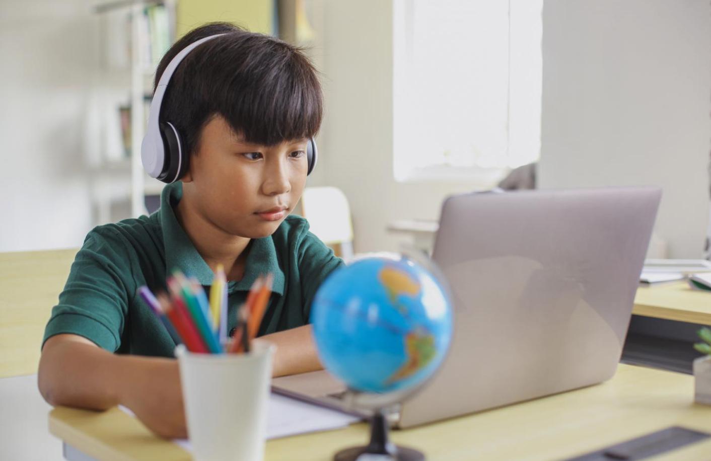 Child wearing dark green polo shirt and headphones, looking at something on a laptop screen open in front of him.