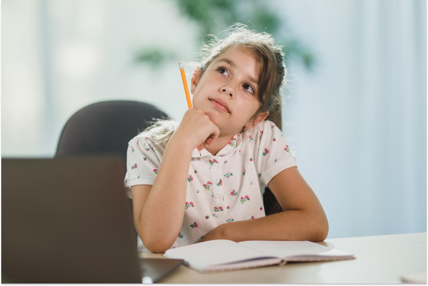 Girl sitting at a table in front of an open notebook. She's holding a pencil and has got her head in her hand, looking up and thinking.