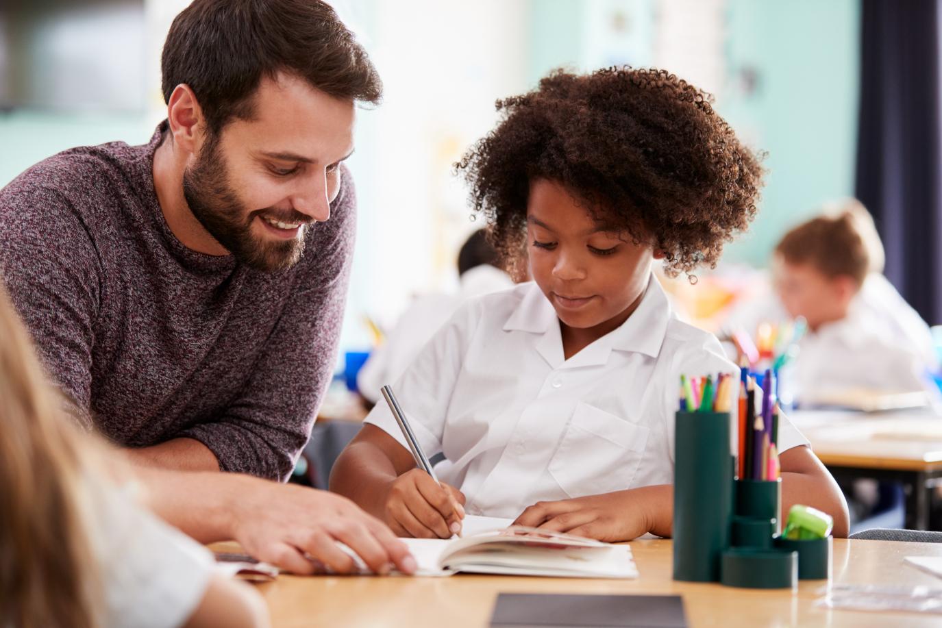 Young smiling male teacher sitting at a classroom table with a young schoolgirl who is writing in a notebook