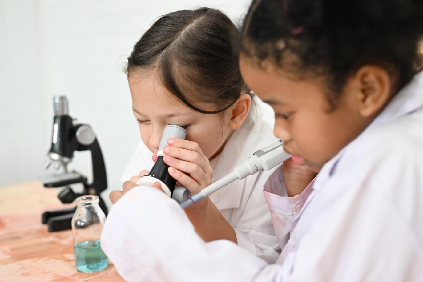 Two young schoolgirls wearing labcoats looking through a microscope in a school science lab