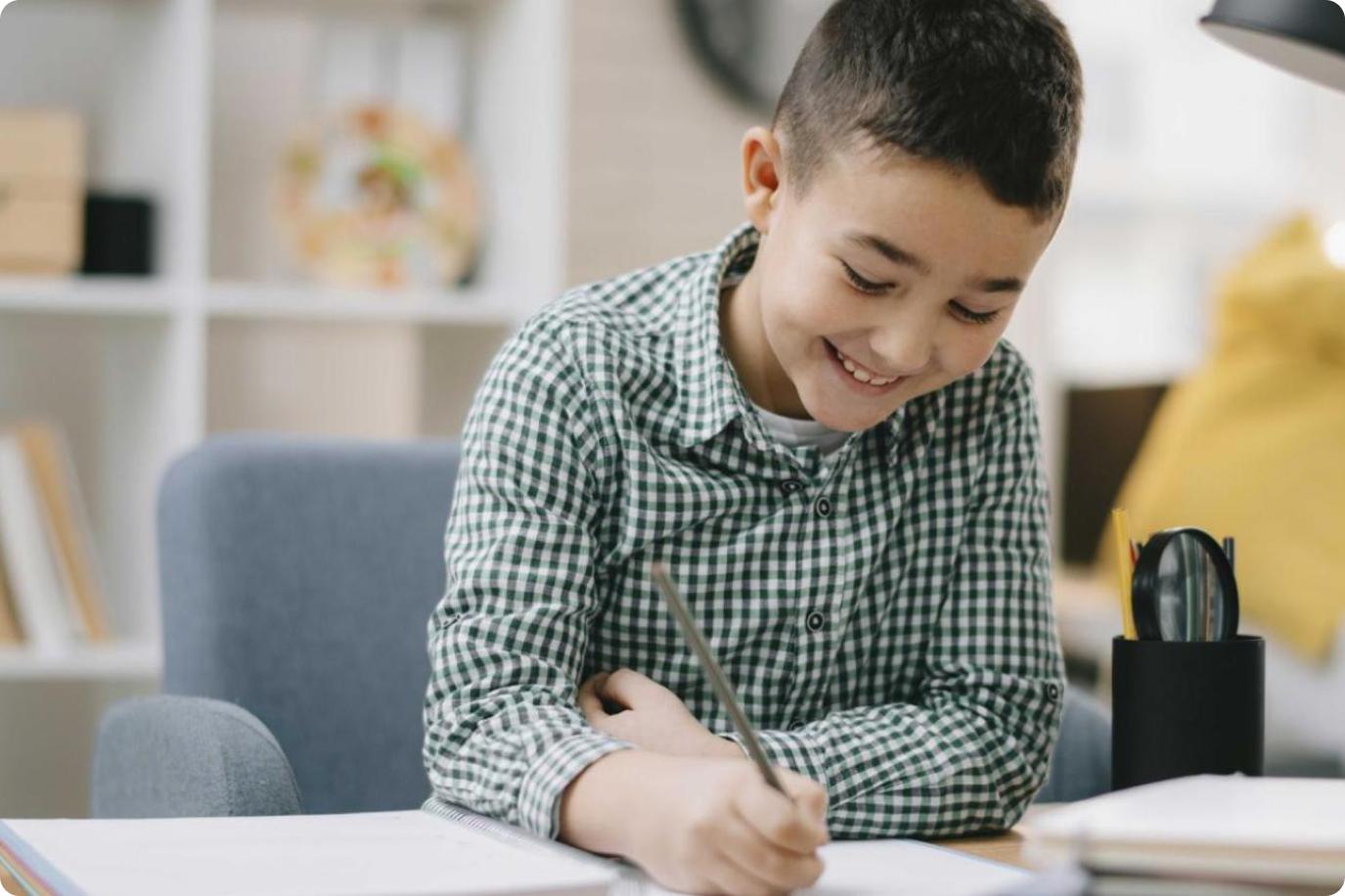 Boy wearing a green checked shirt and grinning while writing in a notebook