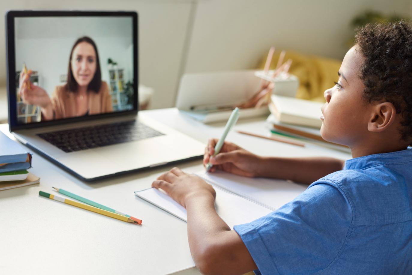 Child writing while listening to an online tutor via a laptop