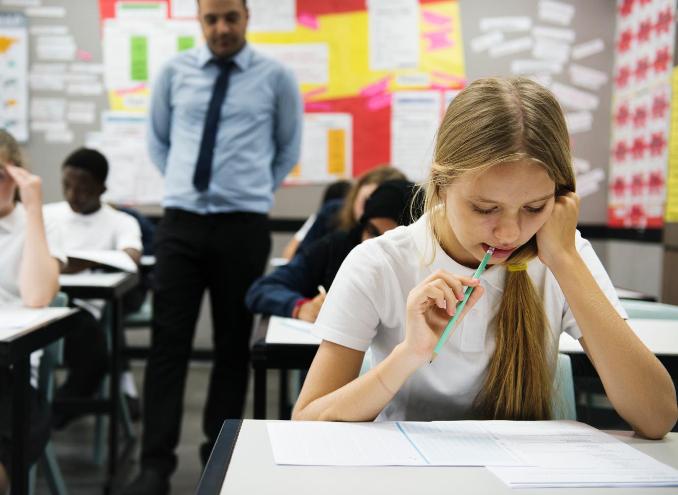 Schoolgirl in white polo top chewing the end of a green pencil while looking at exam sheets on a desk in front of her. In the background, a teacher is walking up the aisle between students' desks. 